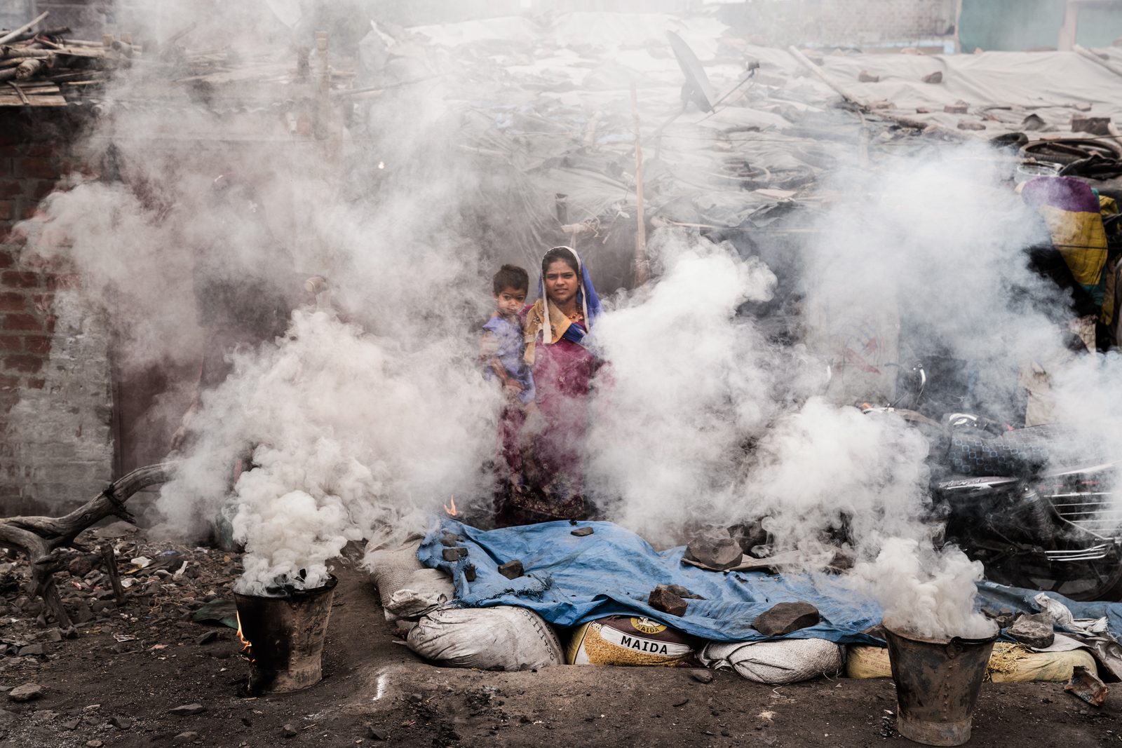 A mother and son wait for their morning chula to fire up while covered in smoke outside their home in Madhya Pradesh, India