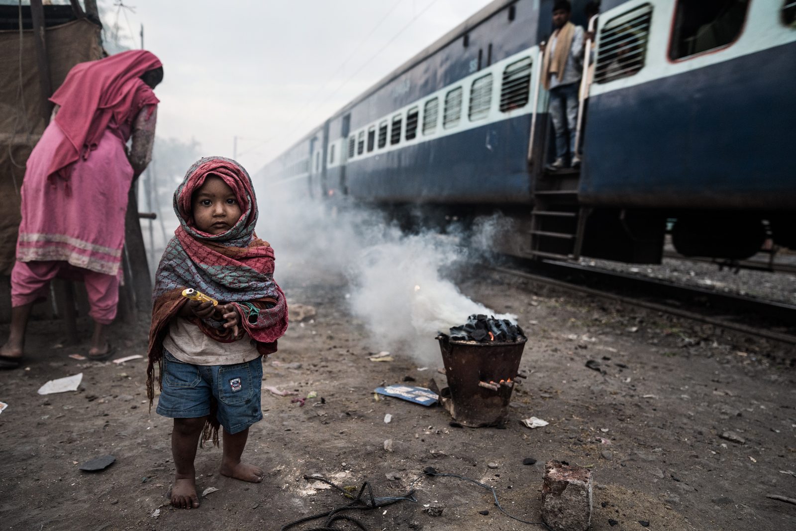 A young boy waits for his family chula to fire up as a train passes by his home in Madhya Pradesh, India
